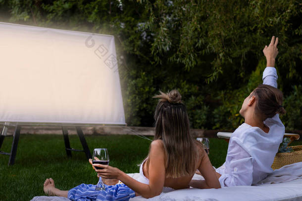 Happy unrecognizable couple sitting in the grass watching a movie in the garden holding red wine gla