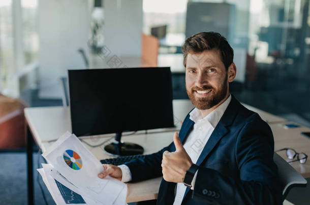 Portrait of <strong>happy</strong> businessman showing thumb up and smiling, sitting near blank empty computer monito