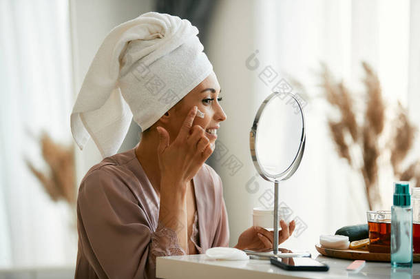 Young happy woman looking herself in a mirror and using moisturizer on her face skin at home.
