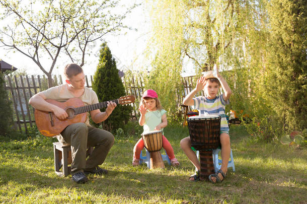 快乐的家庭<strong>爸爸</strong>和两个孩子在户外一起玩乐器。<strong>爸爸</strong>弹吉他，孩子们玩民族鼓。隔离。音乐概念