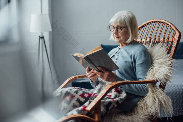 戴眼镜的高级妇女坐在柳条<strong>摇椅</strong>上, 在家看书