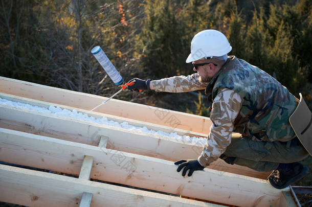 男性建筑工人在木制框架结构房顶上进行保温.男工在未来别墅的屋顶喷涂聚氨酯泡沫。建筑和绝缘概念.