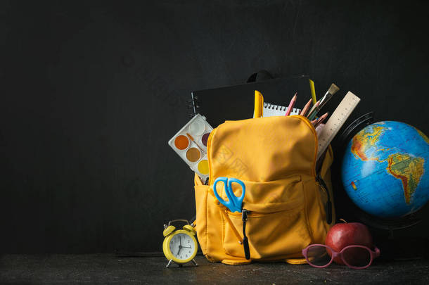 Yellow backpack with school supplies next to the globe, red apple and glasses on the black school bo