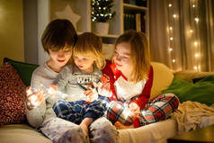 圣诞节前夕，两个大姐姐和她们蹒跚学步的弟弟在一个舒适的客厅里玩圣诞灯。孩子们在寒假期间都呆在家里。带着家人和孩子的冬夜.