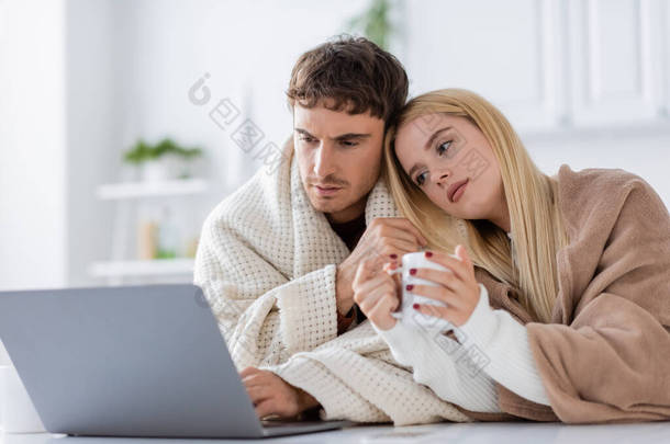 年轻的金发女人披着毛毯，靠着男朋友的肩膀，用笔记本电脑在家里工作