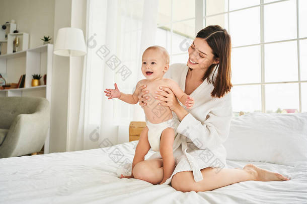 年轻的白人母亲穿着尿布在卧室的床上和她刚出生的儿子玩耍。淋浴后的概念、<strong>婴儿护理</strong>、母性.