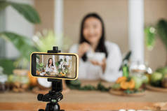 穿着白色浴衣的年轻亚洲女性博客作者或心满意足的创作者坐在木制桌旁，用各种配料在家里准备天然化妆品，一边在碗里搅拌，一边准备奶油.