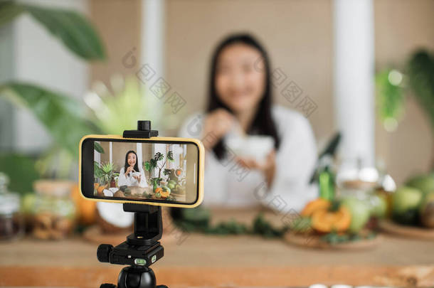 穿着白色浴衣的年轻亚洲女性博客作者或心满意足的创作者坐在木制桌旁，用各种配料在家里准备天然化妆品，一边在碗里搅拌，一边准备奶油.