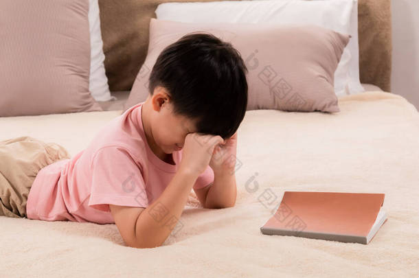 亚洲小男孩在床上看书时感到困倦，揉揉眼睛；小学生在放学后躺在床上看书，做作业时感到很疲倦.