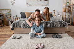 积极的孩子在快乐的父母床边的拖鞋旁边坐在地毯上