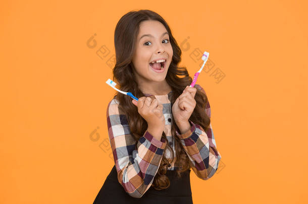 再快两倍 小女孩拿着两把牙刷。 童年时代的女孩聪明的孩子快乐的脸蛋在乎卫生. 刷牙的概念。 牙齿卫生。 女孩可爱的长发拿着牙刷黄色背景.
