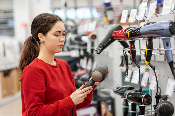 Caucasian young woman with long hair, holding a hair dryer. In the background, shelves with househol