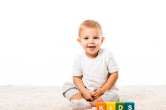 笑的幼儿男孩坐在地毯上附近的彩色立方体与信件孤立在白色