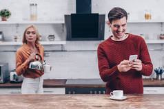 嫉妒的年轻女子与水壶看着微笑的丈夫使用智能手机在厨房 