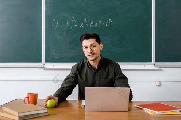 坐在<strong>电脑</strong>桌前、拿着<strong>苹果</strong>、在教室里看相机的男老师