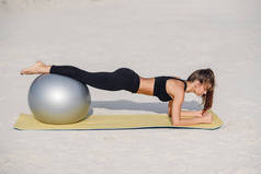 年轻漂亮的健身女孩做俯卧撑运动与健身球在海滩上。体育与健康生活方式概念.