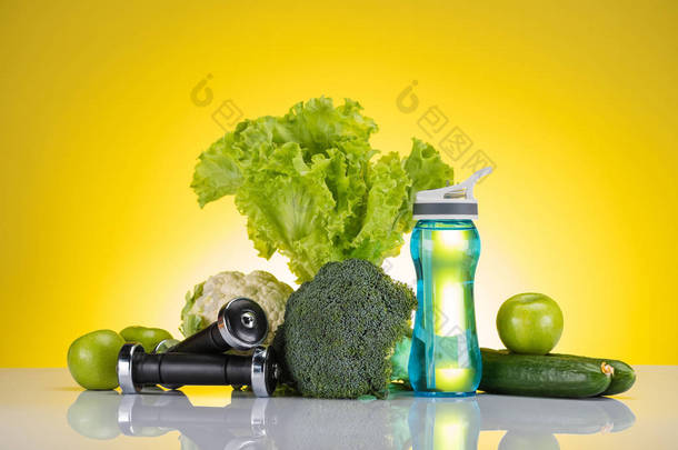 鲜绿色苹果和蔬菜, 哑铃和水瓶在黄色的特写视图