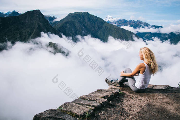 正值年轻的金发女郎穿着积极的穿着坐在洛基山和欣赏惊险的风景高绿色的山覆盖在南美惊人的旅程中的白色雾