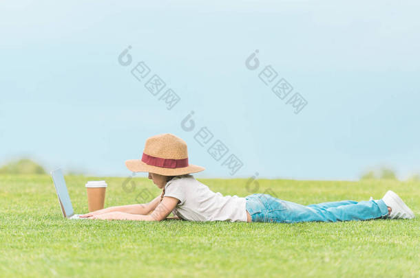 在绿色草地上使用膝上型电脑的可爱儿童的侧面视图