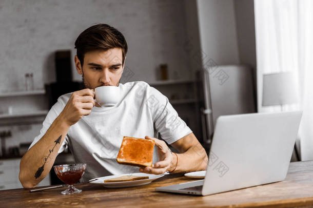 英俊的年轻自由职业者吃烤面包与果酱和咖啡, 看着笔记本电脑屏幕在家里