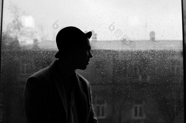 帽子剪影在雨水落在窗口和城市视图背景的白人帅哥的肖像
