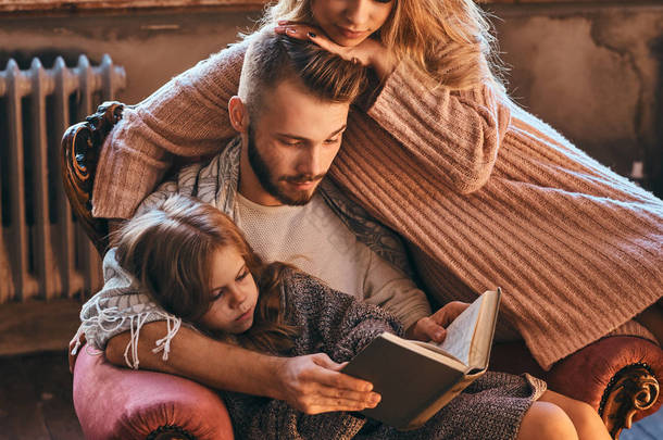 家庭团聚。妈妈, 爸爸和女儿一起看故事书, 坐在沙发上。家庭和父母的概念.