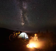年轻女子游客在夜间露营, 在燃烧的篝火旁, 照亮的旅游帐篷, 山地自行车在惊人的美丽的夜空充满星星和银河。天文摄影