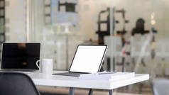 用空白屏幕平板电脑、笔记本电脑、办公用品和咖啡杯在有模糊办公室的白桌子上裁剪的工作场所照片 
