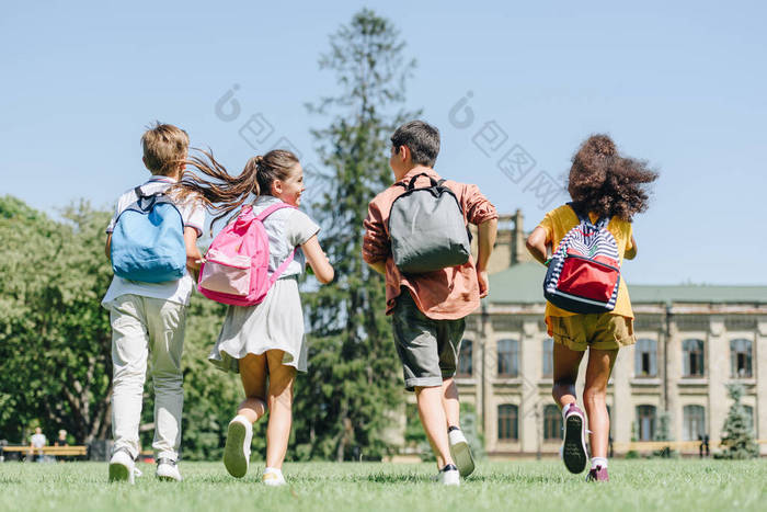 四个多民族学童背着背包在公园的草坪上奔跑的背景