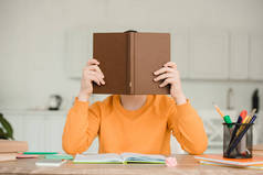 男孩坐在书桌前，在家做功课时，把脸藏在书后面