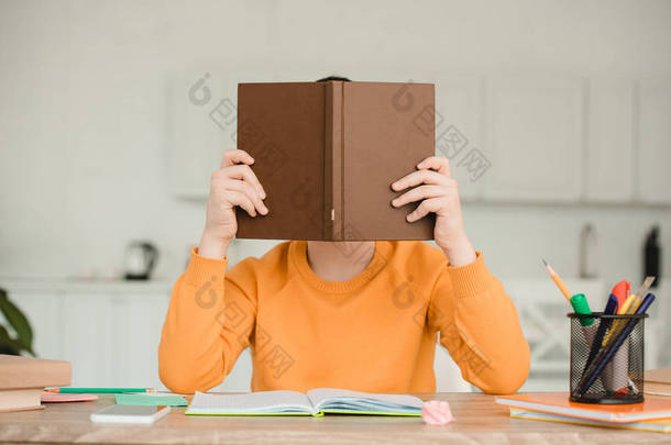 男孩坐在书桌前，<strong>在家</strong>做功课时，把脸藏在书后面