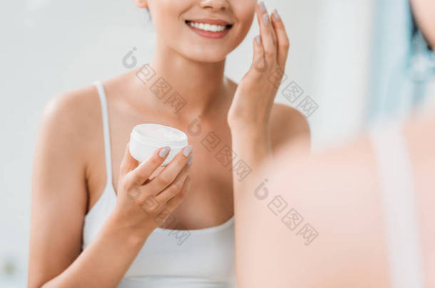 手持容器的微笑女孩裁剪镜头及在浴室镜中涂抹面霜