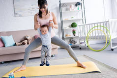 年轻的运动的母亲抱着小孩子, 而站在瑜伽垫在家