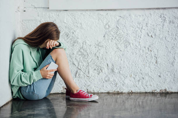 悲伤的哭泣的女孩坐在纹理墙附近，拿着智能手机