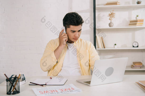 细心的亚洲男子在智能手机上交谈,一边坐在家里的办公桌前,一边使用笔记本电脑