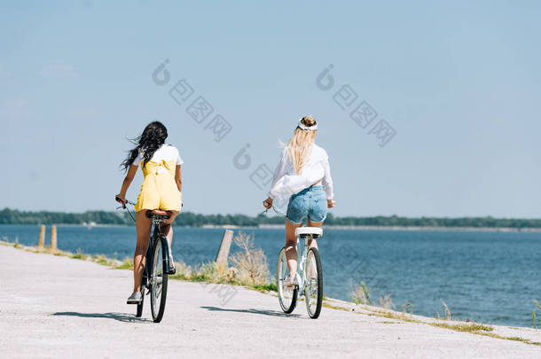 背部的金发和黑发女孩骑自行车在河附近在夏季的背景