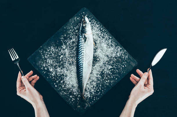 石板板上新鲜生鲭鱼的顶部和人的手用叉子和刀子隔绝在黑色  
