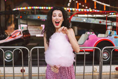 美丽的情感女孩与深色卷曲的头发在礼服站立与粉红色棉花糖在手和惊异看在摄像机在游乐园
