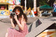 美丽的女士与深色卷曲的头发在太阳镜和帽子坐在手里的小相机和花时间在游乐园与景点背景