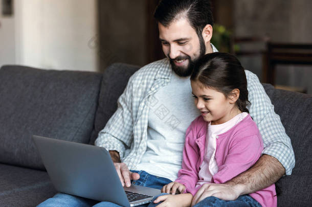 快乐的父亲与女儿坐在沙发上, 使用笔记本电脑