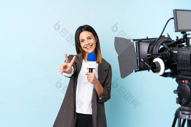 女记者手持话筒，在孤立的蓝色背景上微笑着报道新闻，并展示胜利的标志