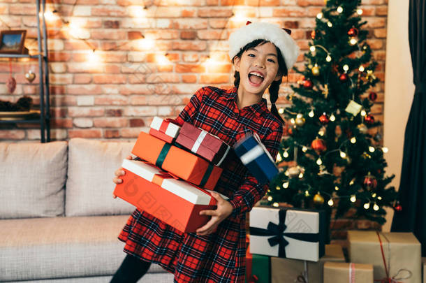 可爱的女孩拿着礼物显示大露齿微笑。可爱的女儿, 圣诞老人的帽子站在圣诞树前。在圣诞节快乐地得到礼物在家里.