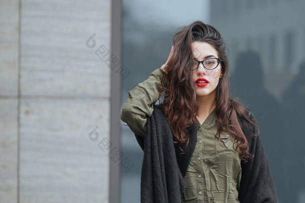 时尚概念︰ 美丽年轻的女孩，长头发，眼镜，红红的嘴唇，站在现代墙身穿绿色套装，灰色的牛仔裤在附近。她想谈电话. 