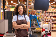 漂亮的年轻微笑着的非洲裔美国女售货员，她的胳膊交叉在胸前，在工作期间用收银机看着你