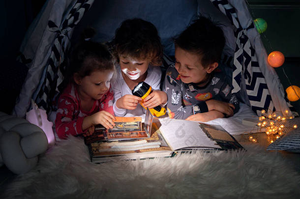 三个孩子<strong>晚上</strong>坐在帐篷里，借着手电筒的光芒阅读一本关于