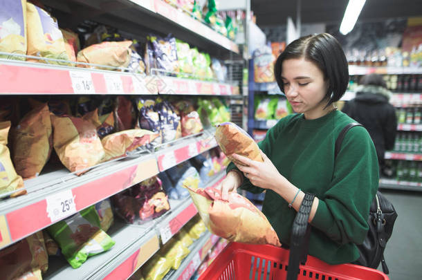 一个小女孩在超级市场买零食。这个女孩在超市里<strong>选择</strong>薯条, 把它们放在一个红色的购物车里。在超级市场购物.