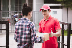 年轻的后勤职业概念。快乐送货的人把他的包给客户在家里。在真正的房子。二十岁代初, 亚洲华人身穿红色马球衫和红帽牛仔裤.