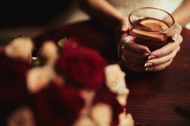 年轻的新娘喝茶。婚礼花束在餐厅的木桌上。新娘拿着一杯茶与拉蒙的裁剪图像