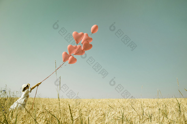 Young romantic girl with red heart balloons walking in a field o