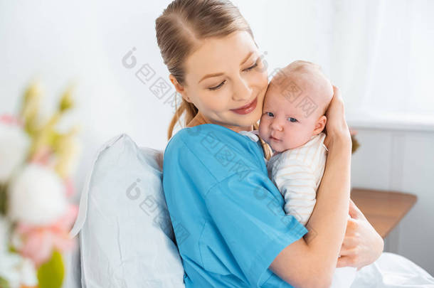 快乐的年轻母亲坐在<strong>病床</strong>上拥抱可爱的婴儿 